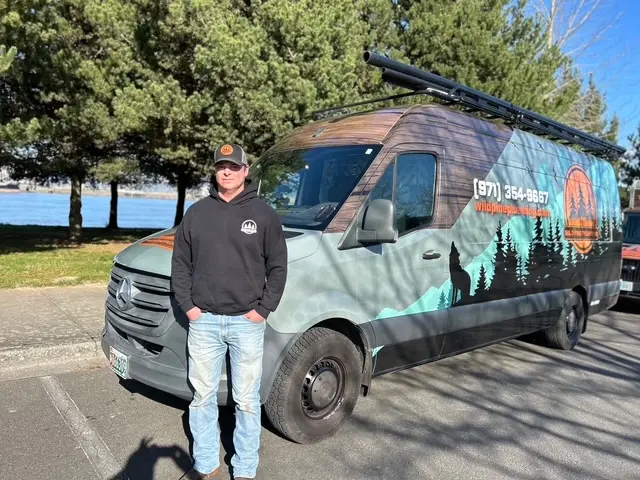 Wild Pine Plumbing owner in branded hoodie standing in front of the wrapped Sprinter van along the Columbia River in St Helens, Oregon